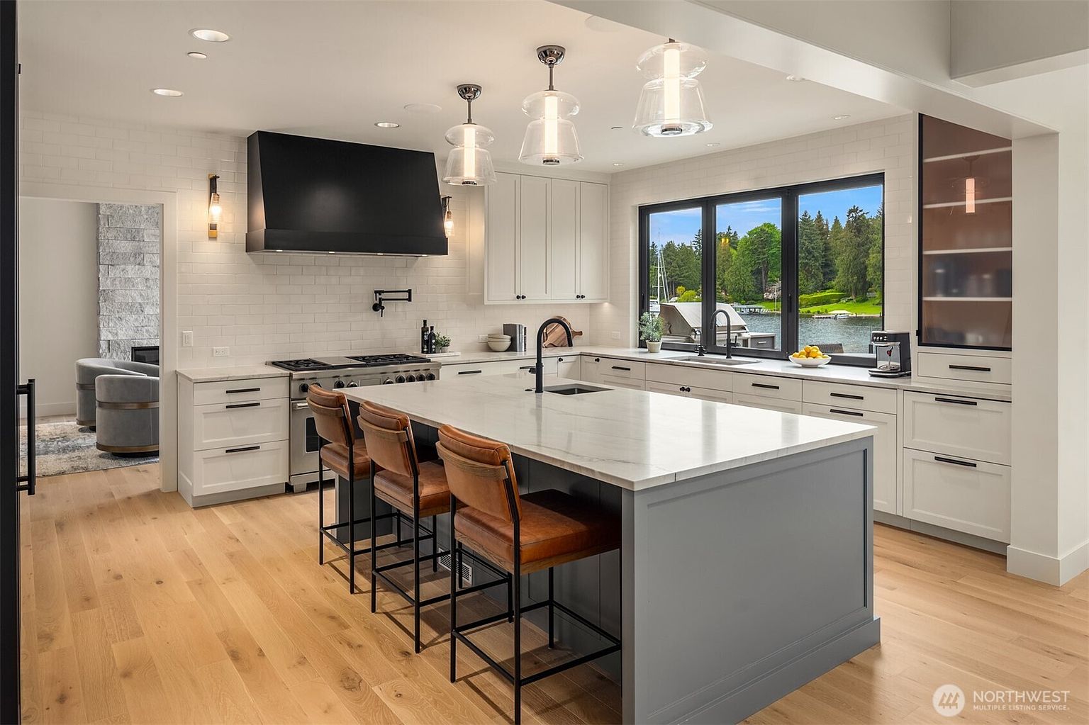 This sophisticated, modern kitchen features a large central island with a grey base and white marble countertop, complemented by three leather-upholstered bar stools. The space is anchored by a striking black range hood, white subway tile backsplash, and light hardwood flooring, all illuminated by elegant glass pendant lights. A large window above the sink provides a serene, picturesque view of the water, creating a bright and airy atmosphere perfect for both cooking and entertaining.