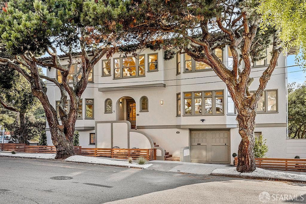 This is a front exterior view of a multi-story home with a light-colored facade and dark-framed windows. Mature trees frame the house, adding to its curb appeal. A paved driveway leads to an attached garage, and a low fence borders the property, enhancing its welcoming appearance.