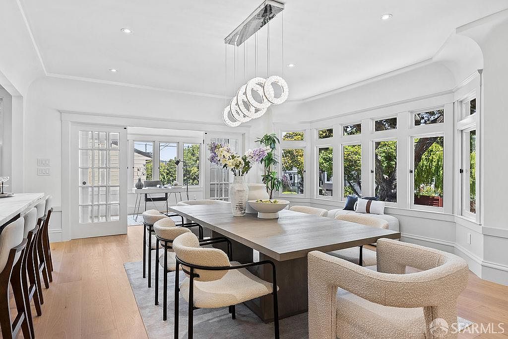 This is a bright and airy dining room featuring a large, modern dining table with seating for eight. The room is well-lit with natural light from the surrounding windows and an elegant chandelier overhead. The decor is minimalist and stylish, creating a sophisticated and inviting atmosphere.