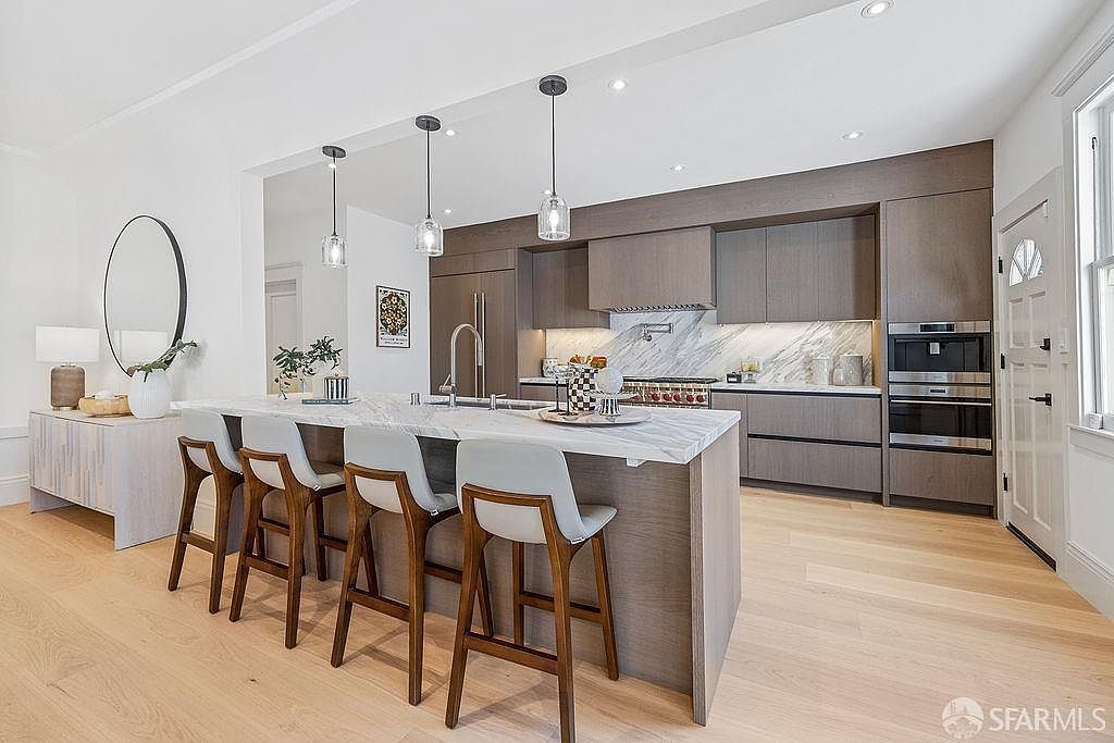 This is a bright and modern kitchen featuring sleek, gray-toned cabinetry and a large island with a white marble countertop and seating for five. Stainless steel appliances are integrated into the cabinetry, and pendant lights hang above the island. The light wood flooring and white walls create a clean and airy atmosphere.