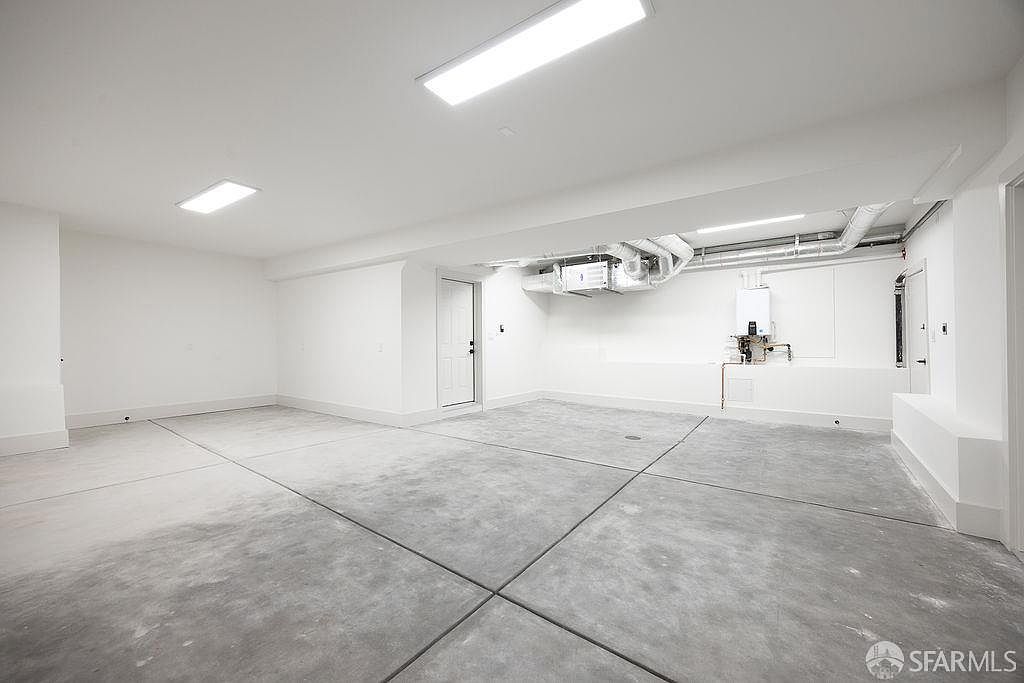 This is a spacious, well-lit garage featuring a concrete floor with expansion joints, white walls, and a white ceiling with recessed lighting. The garage includes utility installations such as a water heater and HVAC system, all neatly arranged. The overall impression is clean and functional, offering ample space for parking and storage.