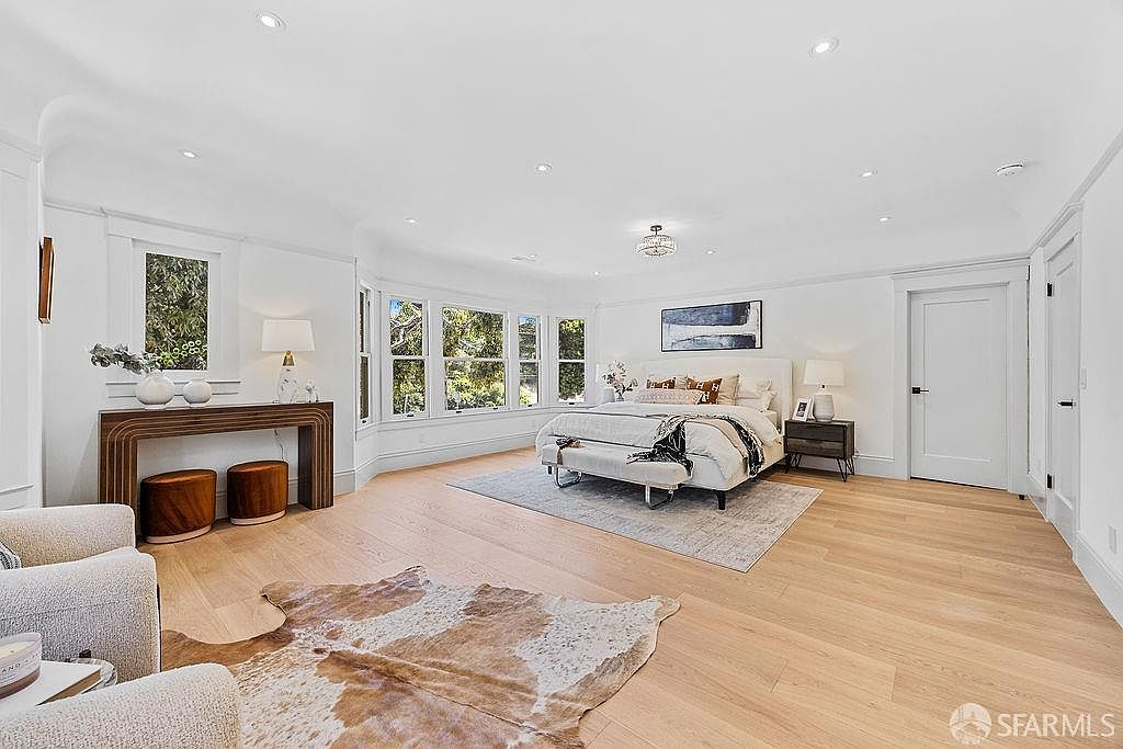This is a bright and spacious primary bedroom featuring hardwood floors, a large bay window, and a neutral color palette. The room is furnished with a king-size bed, a bench, and a side table with a lamp. A console table with decorative items sits against one wall, and a cowhide rug adds texture to the floor. The perspective is from the entrance of the room, showcasing the entire space.
