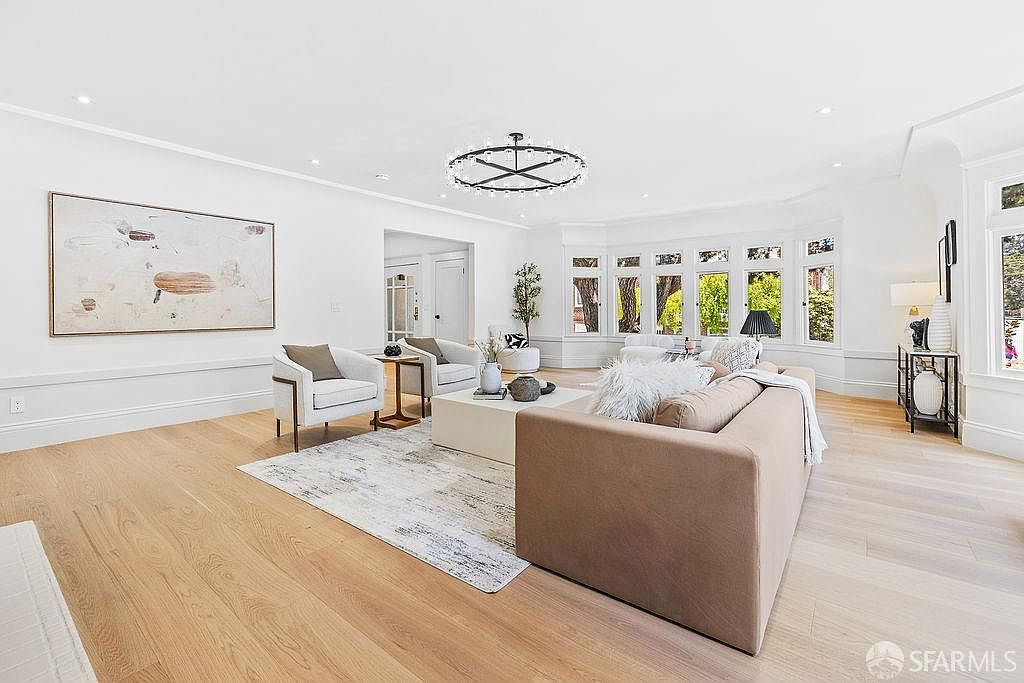 This is an interior shot of a living room featuring a neutral color palette with hardwood floors and white walls. The room is furnished with a beige sofa, two white armchairs, and a light-colored area rug. A modern chandelier hangs from the ceiling, and large windows provide ample natural light, creating a bright and inviting atmosphere.