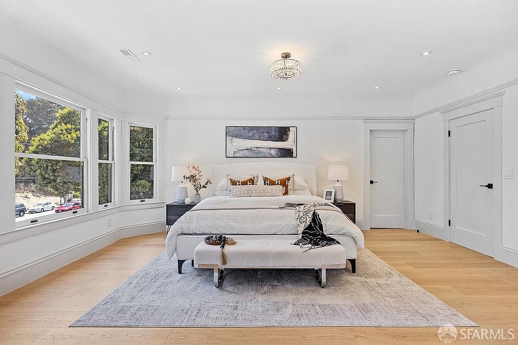 This is a bright and airy primary bedroom featuring a large window with natural light, a neutral color palette, and hardwood floors. The room is centered around a well-appointed bed with decorative pillows and a bench at the foot, complemented by matching nightstands and lamps. An elegant chandelier and artwork add a touch of sophistication.