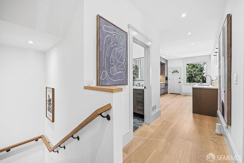 This interior shot showcases a bright hallway and staircase area. The walls are painted white, complemented by light wood flooring and a matching wooden handrail. Artwork adorns the walls, adding a touch of modern elegance to the space.