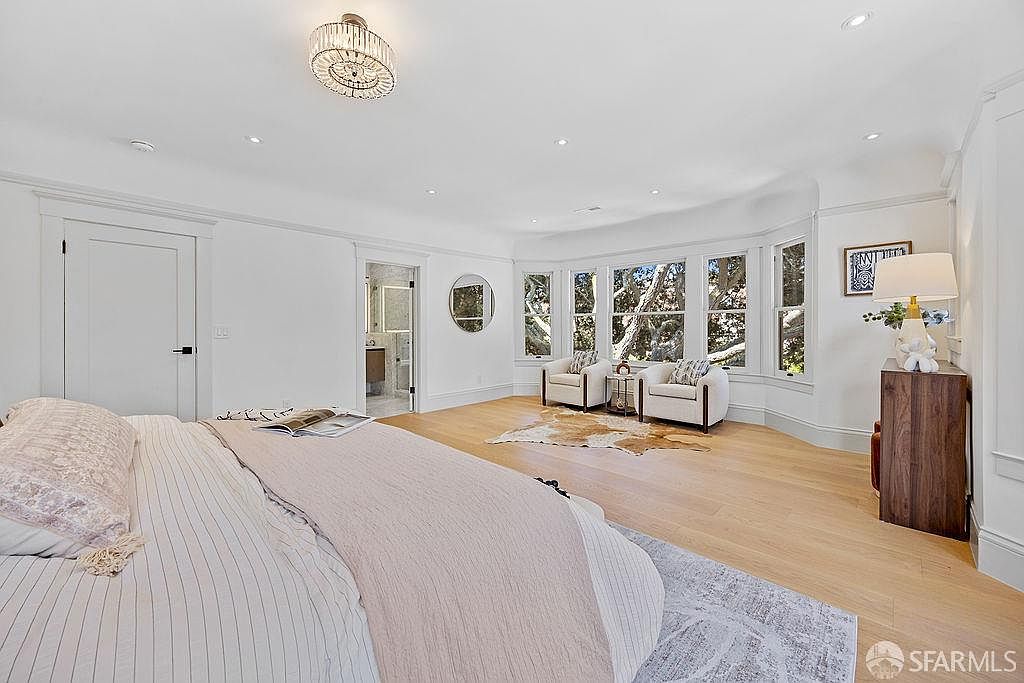 This is a bright and spacious primary bedroom featuring a large bed with light-colored linens, a bay window area with seating, and hardwood floors. The room is painted in neutral tones, creating a serene and inviting atmosphere. A chandelier provides elegant lighting, and a glimpse into an ensuite bathroom adds to the luxurious feel.
