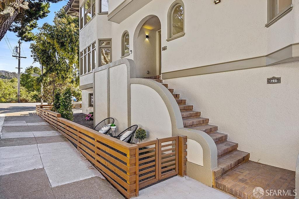 This is a front exterior view of a multi-story home featuring a unique architectural style. The entrance is accessed via brick stairs with curved concrete supports, leading to an arched doorway. A modern wooden fence lines the front of the property, adding privacy and curb appeal.
