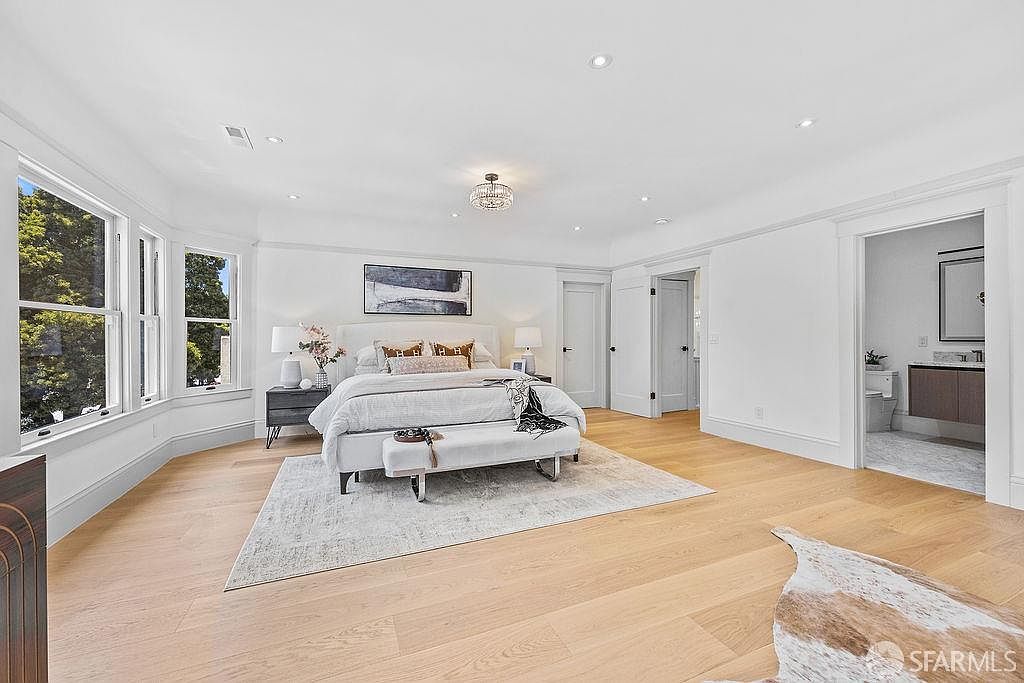 This is a bright and spacious primary bedroom featuring hardwood floors, white walls, and large windows that provide ample natural light. The room is furnished with a king-size bed, a bench at the foot of the bed, and a nightstand with a lamp. A doorway leads to an en-suite bathroom, enhancing the room's appeal as a luxurious and comfortable retreat.