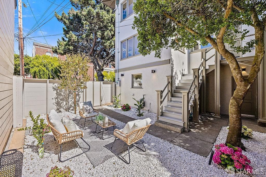 This image showcases a charming outdoor garden area, featuring white gravel ground cover, a small patio with a patterned rug, and comfortable wicker seating. The space is bordered by a white fence and the side of a building with stairs leading to an upper level, creating a cozy and inviting atmosphere. Lush greenery and flowering plants add pops of color, enhancing the garden's appeal.