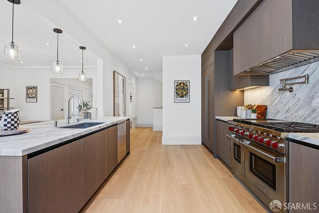 This is a well-lit, modern kitchen featuring sleek, dark wood cabinetry and stainless steel appliances. A large island with a marble countertop dominates the center, complemented by pendant lighting. The kitchen also features a professional-grade range with a stainless steel hood, and the hardwood floors add warmth to the space.