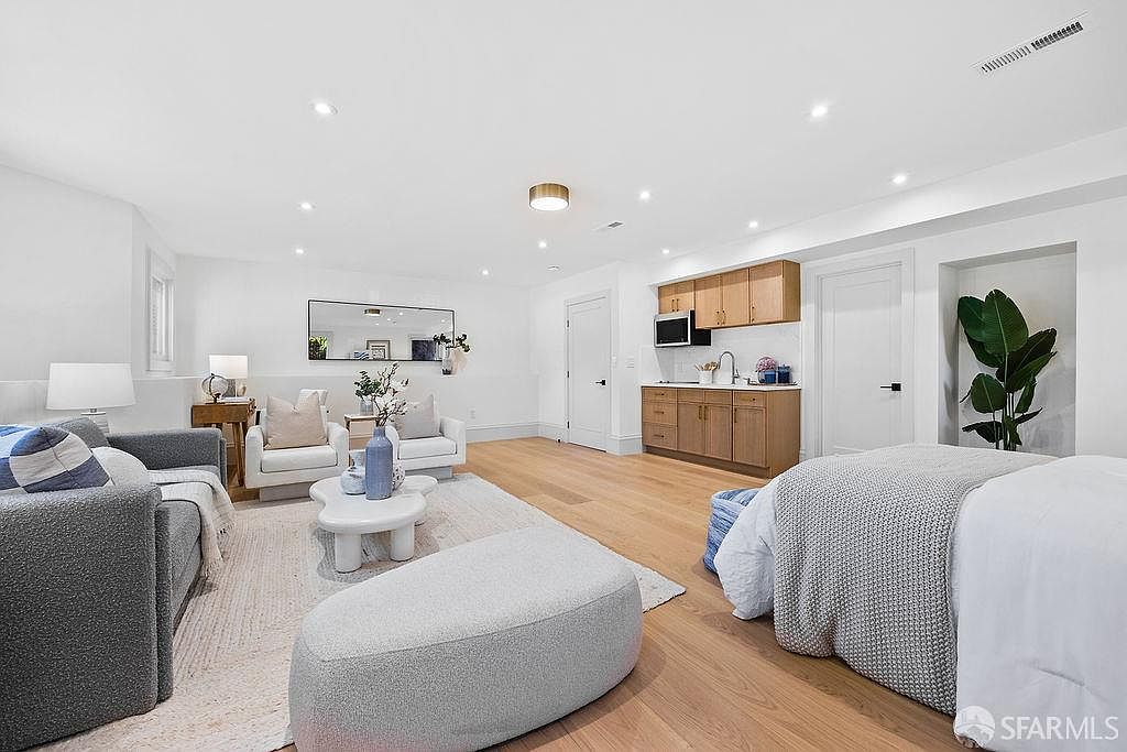 This is a bright and airy living room featuring a modern design with white walls and light wood flooring. The room includes a gray sofa, two white armchairs, a unique coffee table, and a built-in kitchenette with wooden cabinets. A large mirror adds depth to the space, and a potted plant brings a touch of nature indoors.