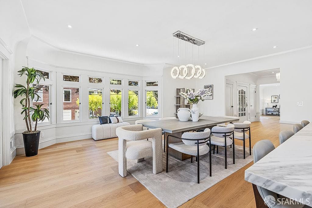 This is an interior shot of a dining room featuring a large dining table with modern chairs, a stylish chandelier, and a bay window with a cushioned bench. The room is bright and airy, with hardwood floors and white walls, creating a sophisticated and inviting atmosphere. A large plant sits in the corner near the bay window.