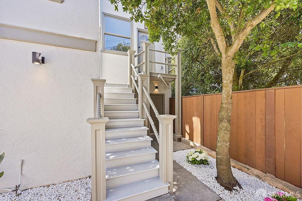 This image showcases an inviting entryway with a staircase leading to an upper level. The stairs feature white risers and treads, complemented by light-colored railings with rope accents. The surrounding landscape includes white gravel, greenery, and a wooden fence, creating a charming and well-maintained exterior.
