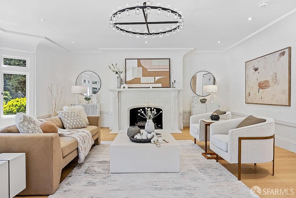 This is a bright and elegant living room featuring a neutral color palette with a tan sofa, two white armchairs, and a large white coffee table centered on a light-colored rug. A decorative fireplace with artwork above serves as a focal point, flanked by round mirrors and lamps on matching cabinets. The room is well-lit by natural light from a window and an ornate chandelier, creating a sophisticated and inviting atmosphere.