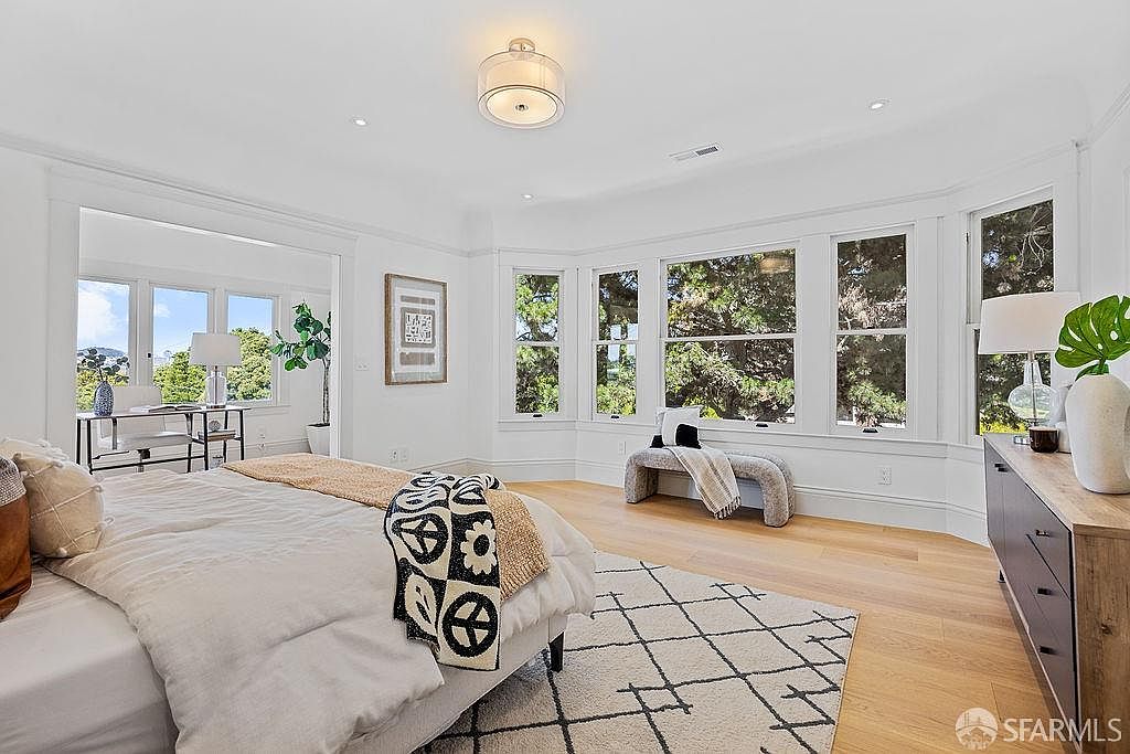 This is a bright and airy primary bedroom featuring a large bay window that provides ample natural light. The room is decorated in a neutral color palette with hardwood floors and a patterned area rug. A comfortable bed is the focal point, complemented by a bench near the window and a dresser with decorative items.
