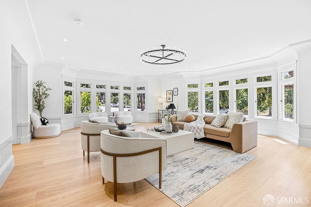 This is a bright and airy living room featuring a neutral color palette, hardwood floors, and large windows that provide ample natural light. The room is furnished with a beige sofa, two armchairs, a white coffee table, and a decorative rug. A modern chandelier hangs from the ceiling, adding a touch of elegance to the space.