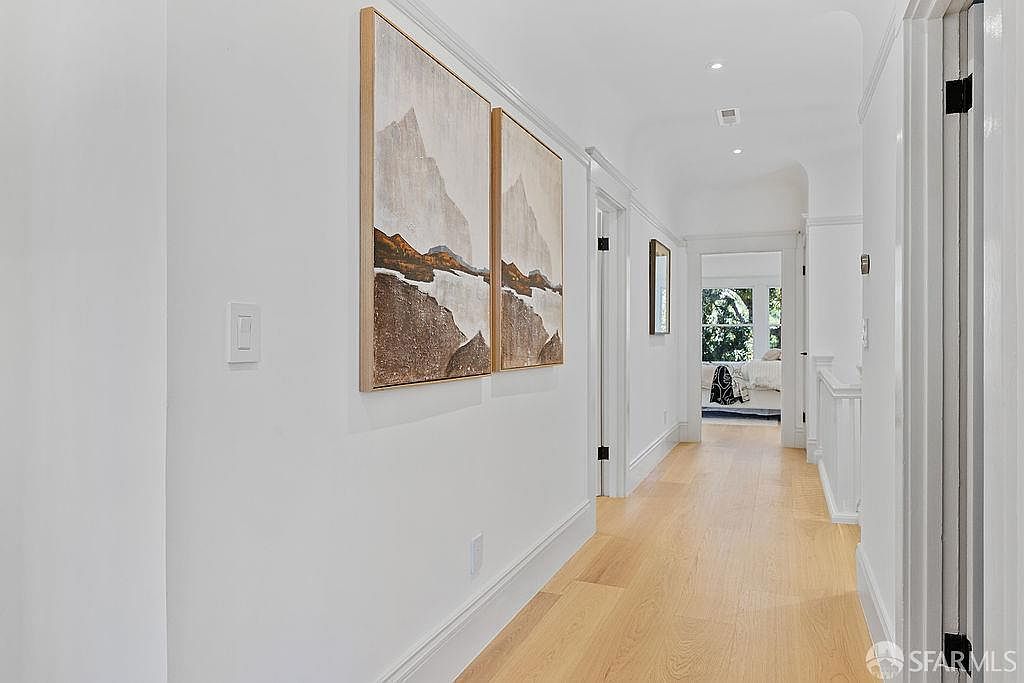 This is an interior shot of a hallway featuring light hardwood floors and white walls. Two framed landscape paintings hang on the left wall, adding a touch of art to the space. The hallway leads to a room with natural light, creating a bright and inviting atmosphere.