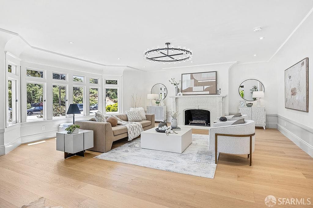 This is a bright and airy living room featuring a large bay window, a neutral-toned sofa, and a modern armchair. A white fireplace with decorative artwork above it serves as a focal point, complemented by a contemporary chandelier. The light hardwood floors and white walls create a spacious and inviting atmosphere.