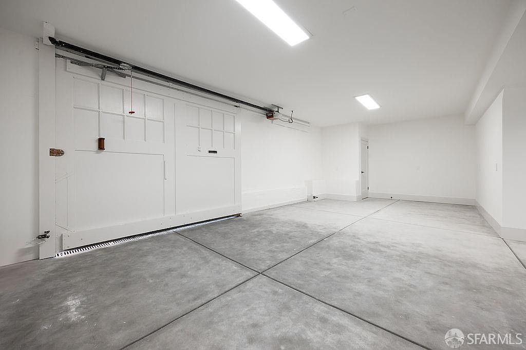 The image showcases a clean and spacious garage interior. The walls and ceiling are painted white, enhancing the brightness, while the floor is concrete with defined sections. A large, white sliding garage door is visible on the left, and overhead lighting illuminates the space, creating a functional and well-maintained area.