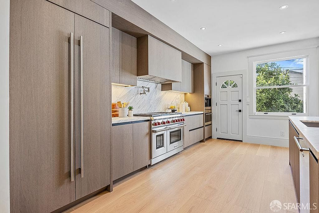 This is a well-lit, modern kitchen featuring sleek, gray-toned cabinetry and stainless steel appliances, including a professional-grade range and a built-in refrigerator. The countertops are light-colored, possibly marble or quartz, and the flooring is light hardwood. A window provides natural light and a view to the outside, while recessed lighting illuminates the space from above. The perspective is from the side, capturing the full length of the kitchen.