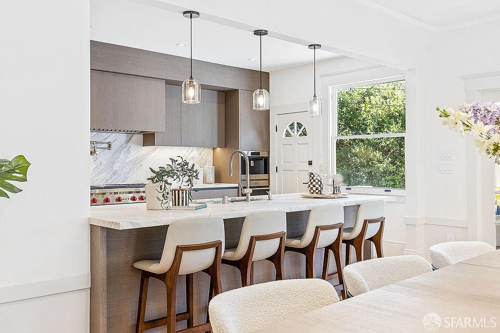 This is a bright and modern kitchen featuring a large island with a white countertop and seating for six. The kitchen includes sleek, gray cabinetry, stainless steel appliances, and pendant lighting. A window provides natural light and a view to the outside, enhancing the inviting atmosphere.