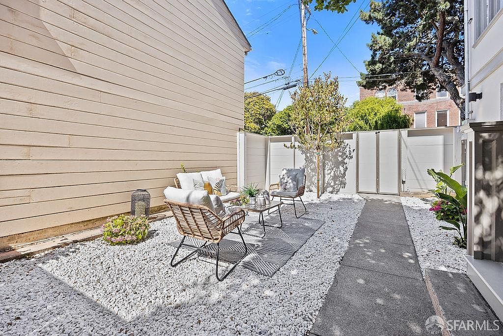 This image showcases a charming outdoor patio area, featuring white gravel ground cover, a paved walkway, and a cozy seating arrangement with wicker chairs and a small table. A light-colored building wall provides a backdrop, while a white fence encloses the space, creating a private and inviting outdoor retreat. The area is decorated with plants and a lantern, adding to the aesthetic appeal.