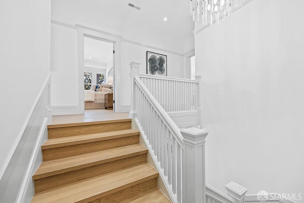 This interior shot showcases a well-lit hallway with wooden stairs leading upwards. The stairs feature a light wood finish and are complemented by a white railing and trim. A doorway at the top of the stairs offers a glimpse into another room, suggesting a spacious and inviting home layout.