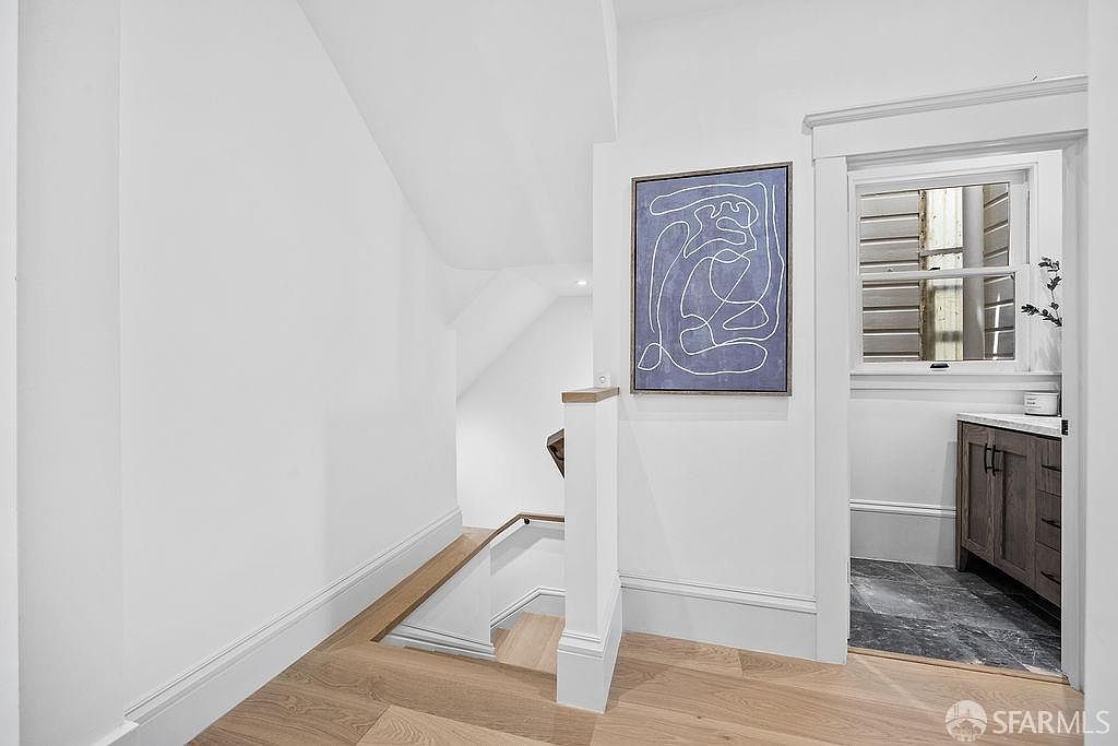 This interior shot showcases a bright hallway with stairs leading to a lower level. The walls are painted white, complemented by light wood flooring. An abstract art piece hangs on the wall, adding a touch of modern elegance, while a glimpse into a bathroom reveals a dark marble floor and a wooden vanity.