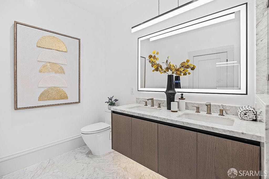 This is a well-lit bathroom featuring a double vanity with a marble countertop and modern faucets. A large, illuminated mirror hangs above the vanity, reflecting the clean, minimalist design. A piece of artwork hangs on the wall next to a toilet, adding a touch of elegance to the space.