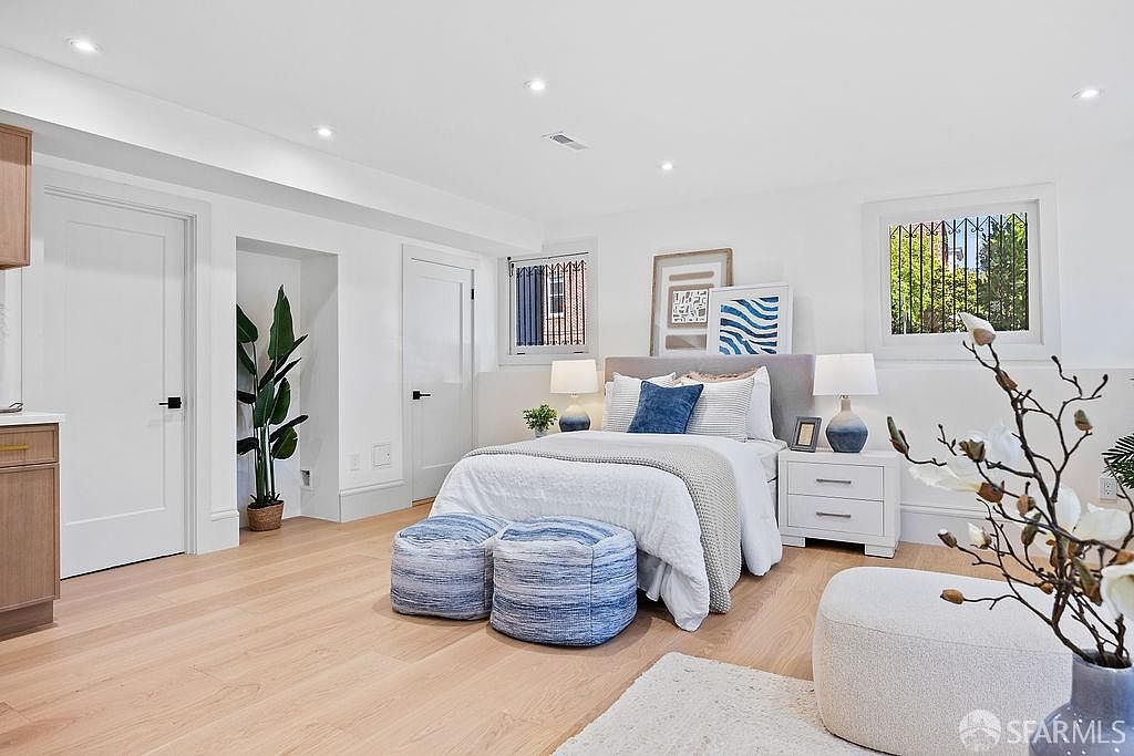 This is a bright and airy primary bedroom featuring light hardwood floors, white walls, and recessed lighting. The bed is neatly made with white linens and blue accents, complemented by matching bedside tables and lamps. Two blue striped ottomans sit at the foot of the bed, adding a touch of color and texture to the space.