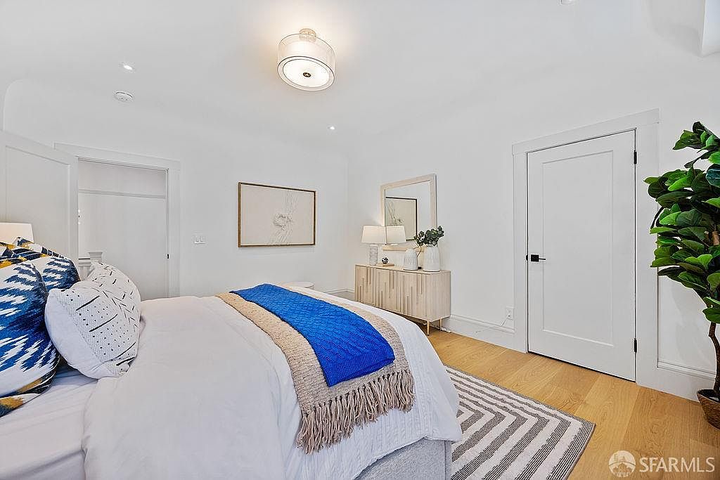 This is a bright and airy primary bedroom featuring white walls, light hardwood floors, and a modern aesthetic. The room includes a bed with decorative pillows and a blue throw, a light wood dresser with lamps and a mirror, and a large potted plant in the corner. A striped rug adds visual interest, and the overall impression is clean and inviting.