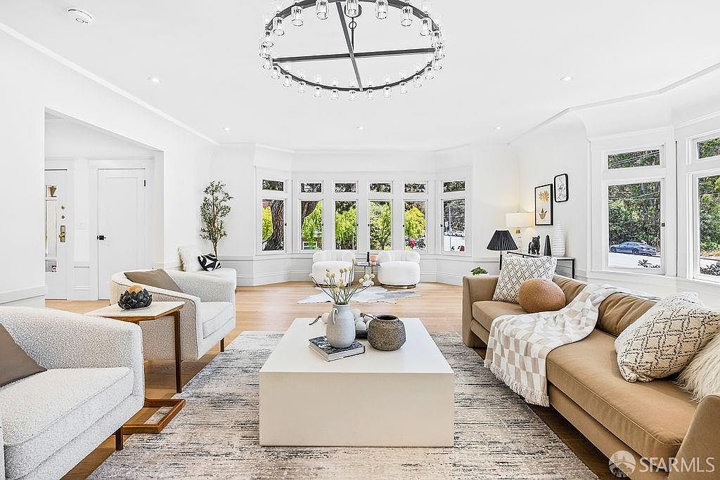 This is a bright and airy living room featuring a large bay window that floods the space with natural light. The room is furnished with a neutral-toned sofa, armchairs, and a large coffee table, creating a comfortable and inviting atmosphere. A modern chandelier hangs from the ceiling, adding a touch of elegance.
