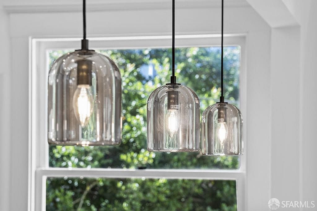 The image showcases three pendant lights with glass shades, suspended against a backdrop of a window offering a view of lush greenery. The lights feature a modern design with a vintage-style bulb, adding a touch of elegance and warmth to the space. The composition highlights the fixtures as a focal point, emphasizing their aesthetic appeal and contribution to the interior's ambiance.