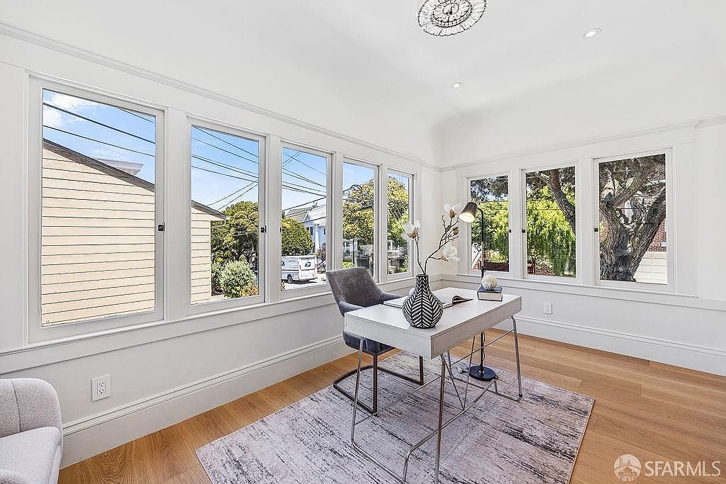 This is a bright and airy home office featuring a white desk, a gray chair, and a large window offering ample natural light. The room is decorated in a minimalist style with light wood floors and a neutral-colored rug. The overall impression is clean, modern, and conducive to productivity.