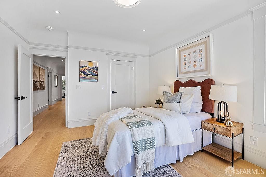 This is a cozy bedroom featuring a bed with a brown headboard, white bedding, and a patterned throw. A wooden nightstand with a lamp sits beside the bed, and artwork adorns the walls. The room has hardwood floors and white walls, creating a bright and inviting atmosphere.