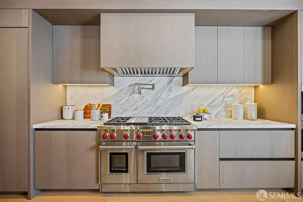 This is a well-lit, modern kitchen featuring light wood cabinetry and stainless steel appliances. The focal point is a Wolf range with a matching hood, set against a marble backsplash. The countertops are light-colored, and under-cabinet lighting adds a warm glow to the space.