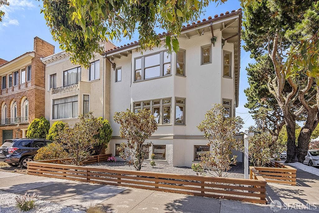 This is a front view of a multi-story residential building with a light-colored facade and a red tile roof. The property features well-maintained landscaping with a wooden fence, small trees, and shrubs. The architectural style appears classic and well-preserved, suggesting a desirable residential property.