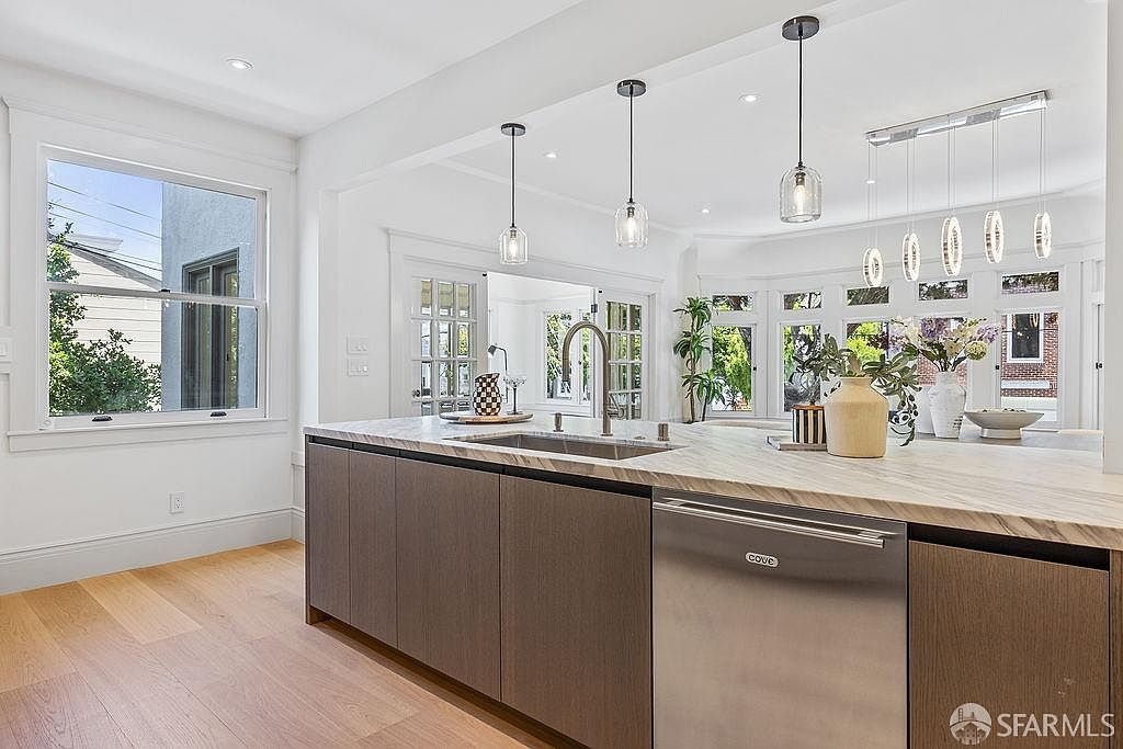 This is a bright and modern kitchen featuring a large island with a marble countertop and a stainless steel sink. The cabinetry is a sleek, dark wood, and stainless steel appliances are integrated seamlessly. Natural light floods the space through large windows, creating an inviting atmosphere.