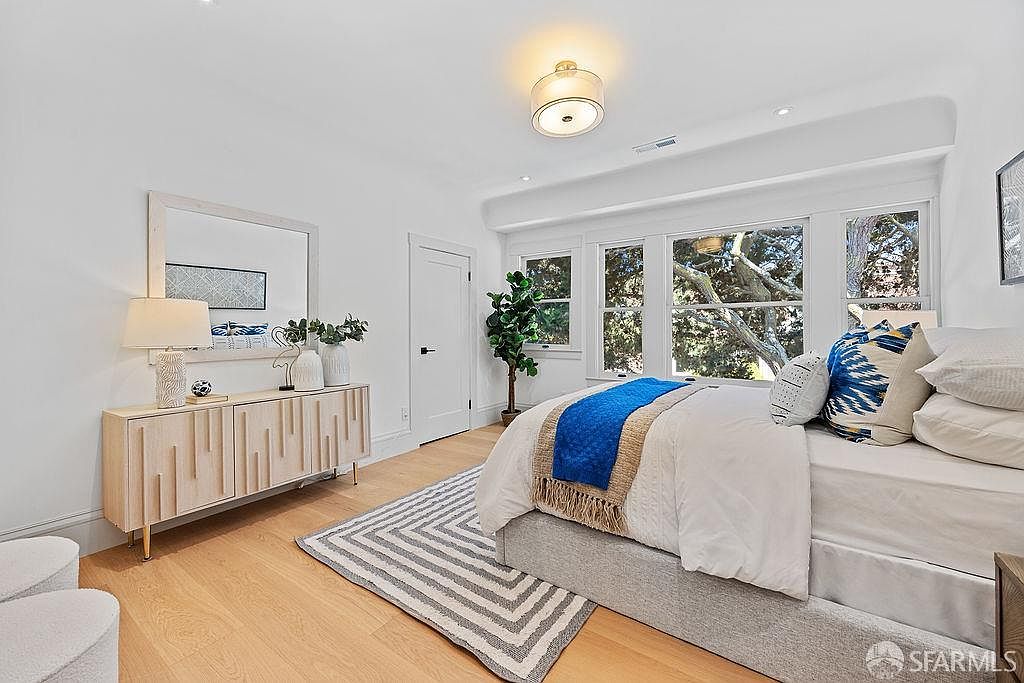 This is a bright and airy primary bedroom featuring a neutral color palette with pops of blue. The room includes a large bed with multiple pillows and a blue throw, a light wood dresser with a mirror above, and a patterned rug. Natural light floods the space through a set of windows, creating a serene and inviting atmosphere.