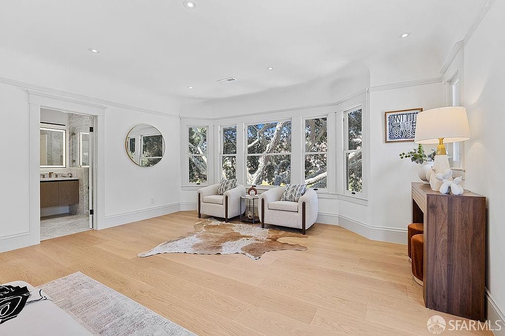 This is a bright and spacious primary bedroom featuring hardwood floors, white walls, and large windows offering natural light. The room includes two armchairs, a console table with a lamp, and a decorative rug, creating a cozy and inviting atmosphere. A glimpse into an ensuite bathroom and a round mirror add to the room's appeal.