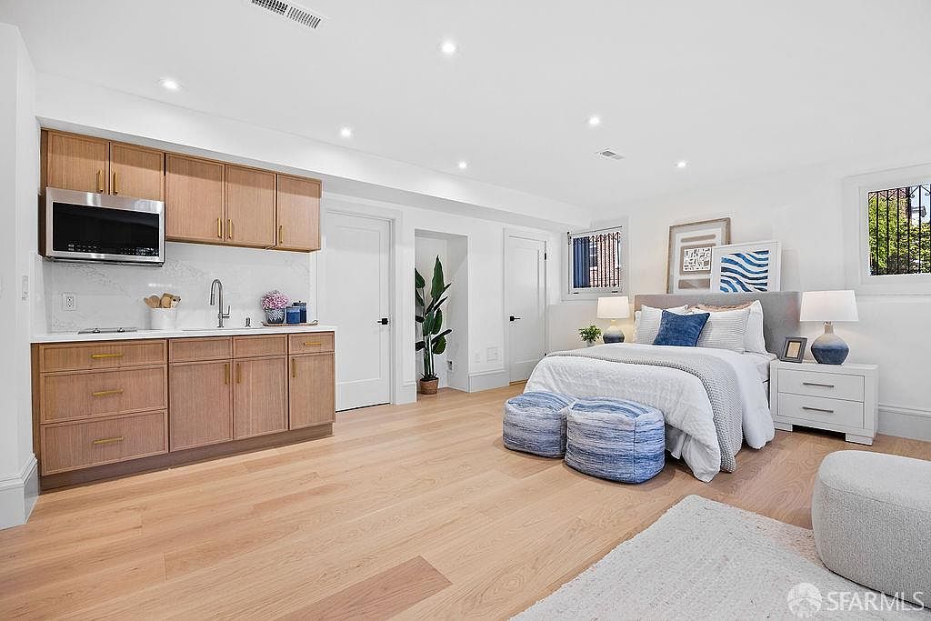 This is a well-lit bedroom featuring a kitchenette area with wooden cabinets and stainless steel appliances. The bedroom area includes a bed with blue accents, matching nightstands, and artwork on the walls. The room has a clean, modern aesthetic with light wood flooring and white walls, creating a bright and inviting space.