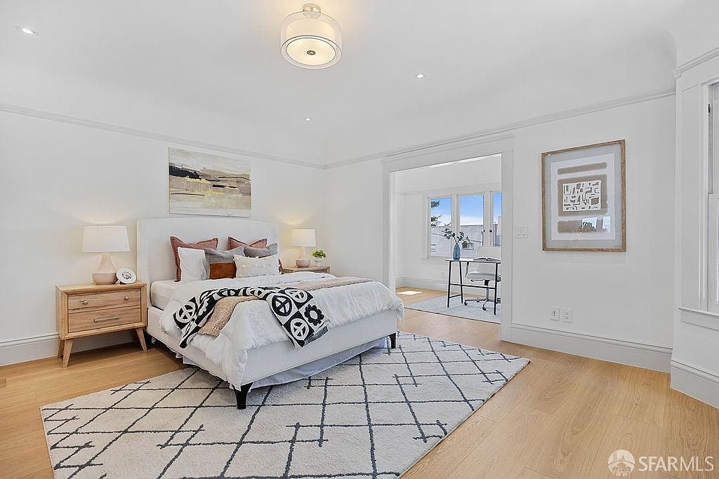 This is a bright and airy primary bedroom featuring a white bed with decorative pillows and a patterned throw. The room has hardwood floors covered by a geometric rug, and a light wood nightstand sits beside the bed. A doorway leads to another room, possibly an office or sitting area, enhancing the sense of space and luxury.