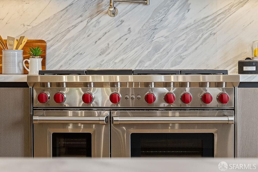 The image showcases a high-end stainless steel range with red knobs, set against a marble backsplash. The appliance features double ovens and a sleek design, suggesting a modern and luxurious kitchen. The focus is on the quality and features of the cooking appliance.