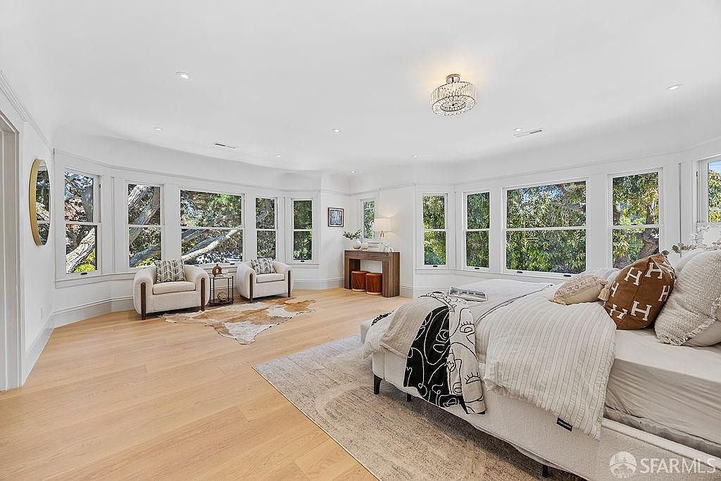 This is a bright and spacious primary bedroom featuring large windows that provide ample natural light and views of the surrounding greenery. The room is tastefully decorated with neutral tones, including a light wood floor, a plush rug, and a comfortable bed with decorative pillows and a throw. The overall impression is one of tranquility and luxury.