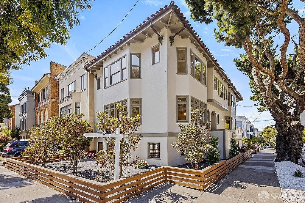 This is a front exterior view of a multi-story house with a well-maintained front yard. The house features a light-colored facade, multiple windows with dark frames, and a red tile roof. A low wooden fence surrounds the yard, which contains small trees and shrubs, enhancing the property's curb appeal.