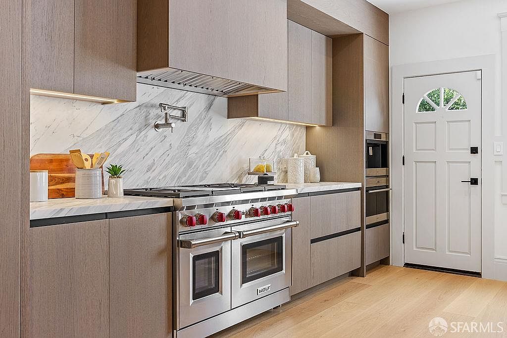 This is a well-lit kitchen featuring modern cabinetry with a wood grain finish, stainless steel appliances including a Wolf range, and marble backsplash. The kitchen has a clean and contemporary design, with a focus on high-end finishes and functionality. A white door with a decorative window is visible on the right.
