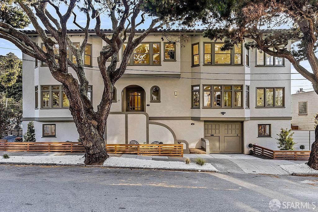 This is a front view of a multi-story home with a light-colored exterior and dark trim around the windows. A large tree partially obscures the facade, adding a natural element to the scene. The property features a well-maintained front yard with white gravel and a modern horizontal slat fence, enhancing its curb appeal.