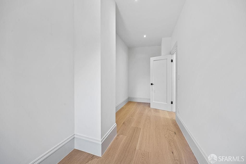 This is an interior shot of a hallway featuring light hardwood flooring and white walls with gray trim. A partially open white door is visible at the end of the hallway, leading into another room. The space is well-lit, creating a clean and modern aesthetic.