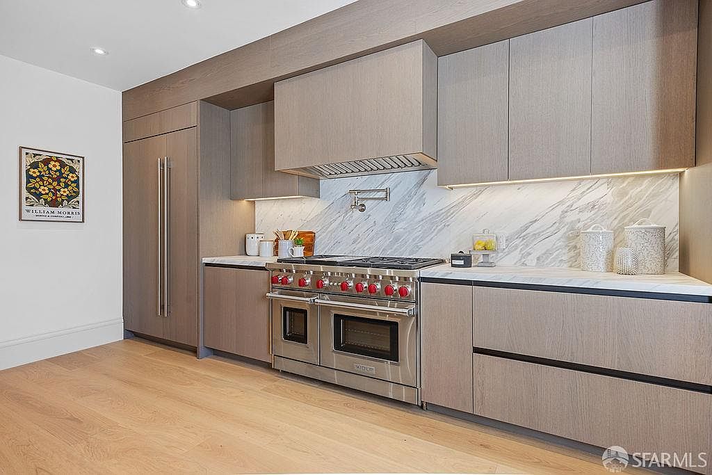 This is a well-lit, modern kitchen featuring light wood cabinetry and stainless steel appliances. The kitchen includes a professional-grade range with red knobs, a marble backsplash, and a built-in refrigerator. The overall impression is clean, sophisticated, and functional, perfect for a home chef.