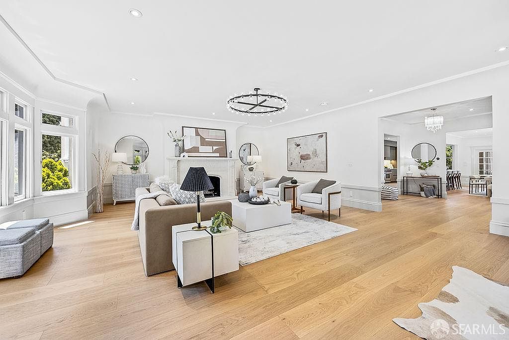 This is a bright and spacious living room featuring hardwood floors, white walls, and elegant architectural details. The room is furnished with a comfortable sofa, armchairs, a modern chandelier, and decorative mirrors, creating a sophisticated and inviting atmosphere. Large windows provide ample natural light, enhancing the room's appeal.